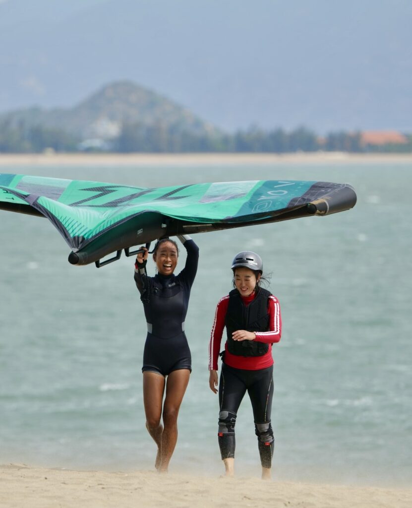 Wing Foil instructor walking on the beach laughing with her student after successfully finishing her wing foil lesson with her rental gear from Jeju Kite Lab. Vietnam winter camp. Wing foil lessons Jeju island South Korea.