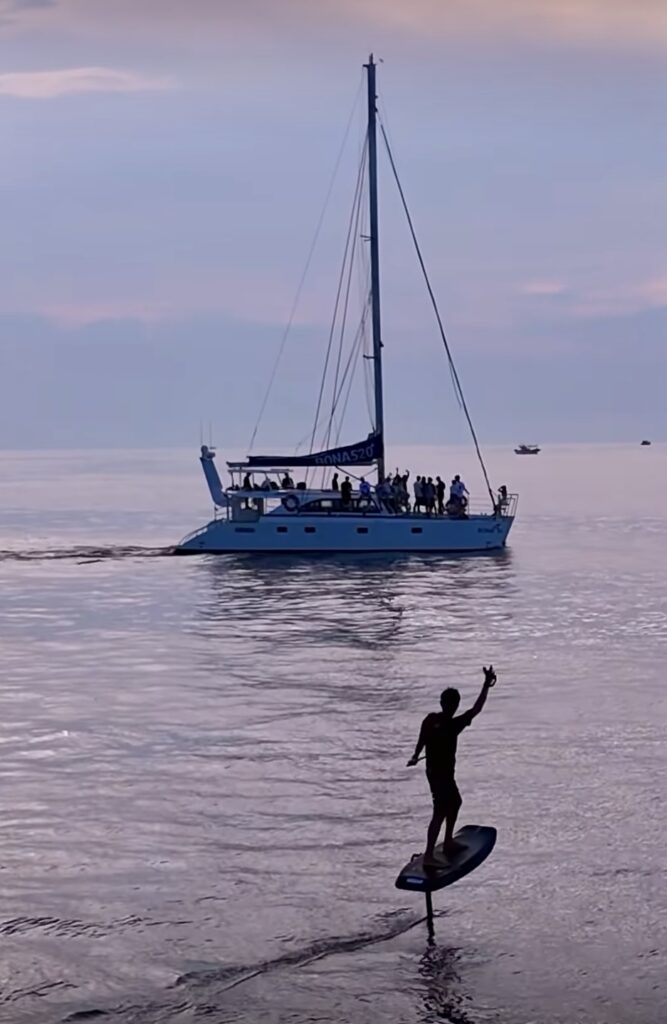 eFoiling in jeju during a sunset waiving to a sailboat while cruising on the water. Enjoying watersports during travels to Jeju South Korea